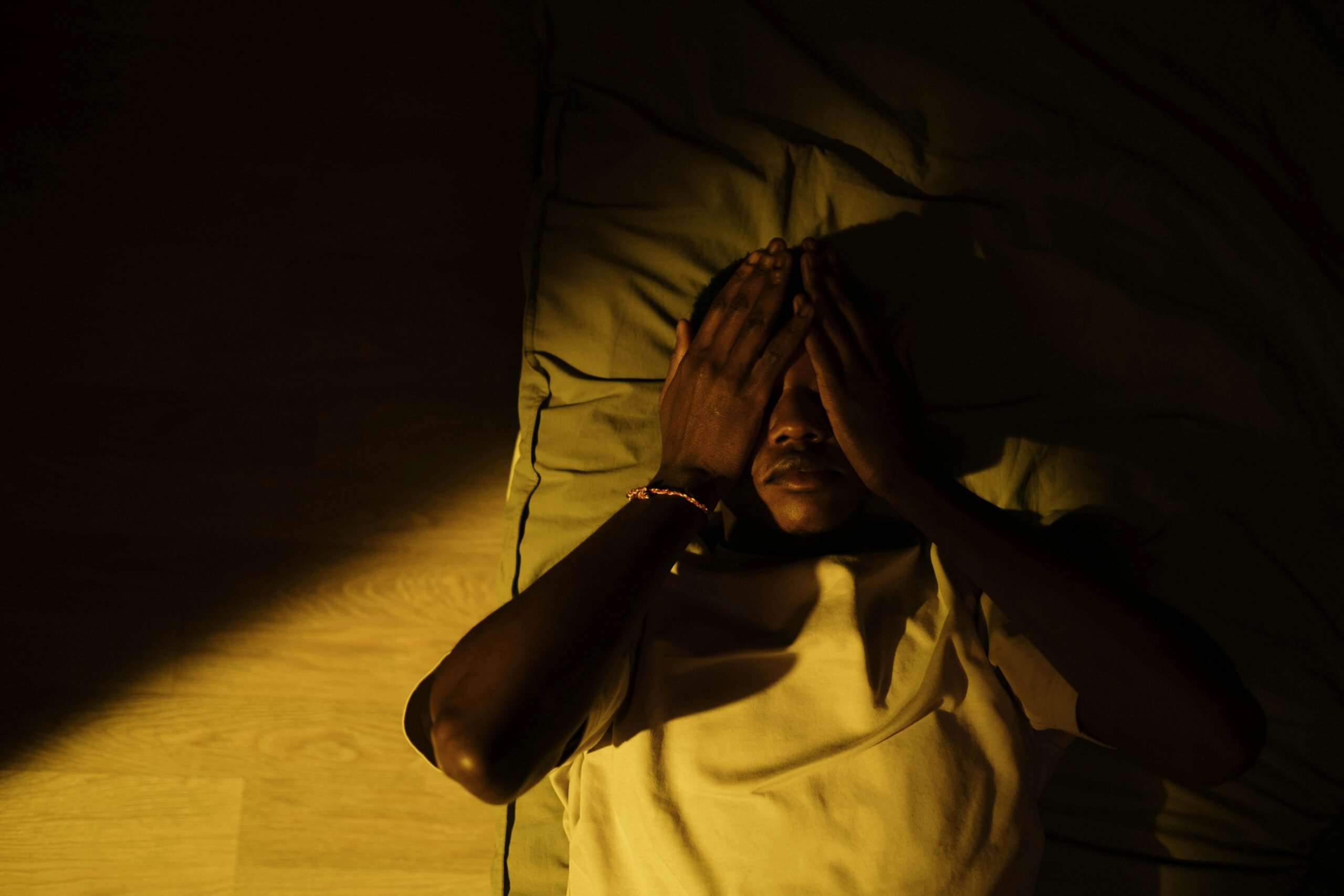 Moody portrait of a man lying down with hands on face in a dimly lit room.